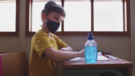 Boy-wearing-face-mask-sanitizing-his-hands-while-sitting-on-his-desk-at-school