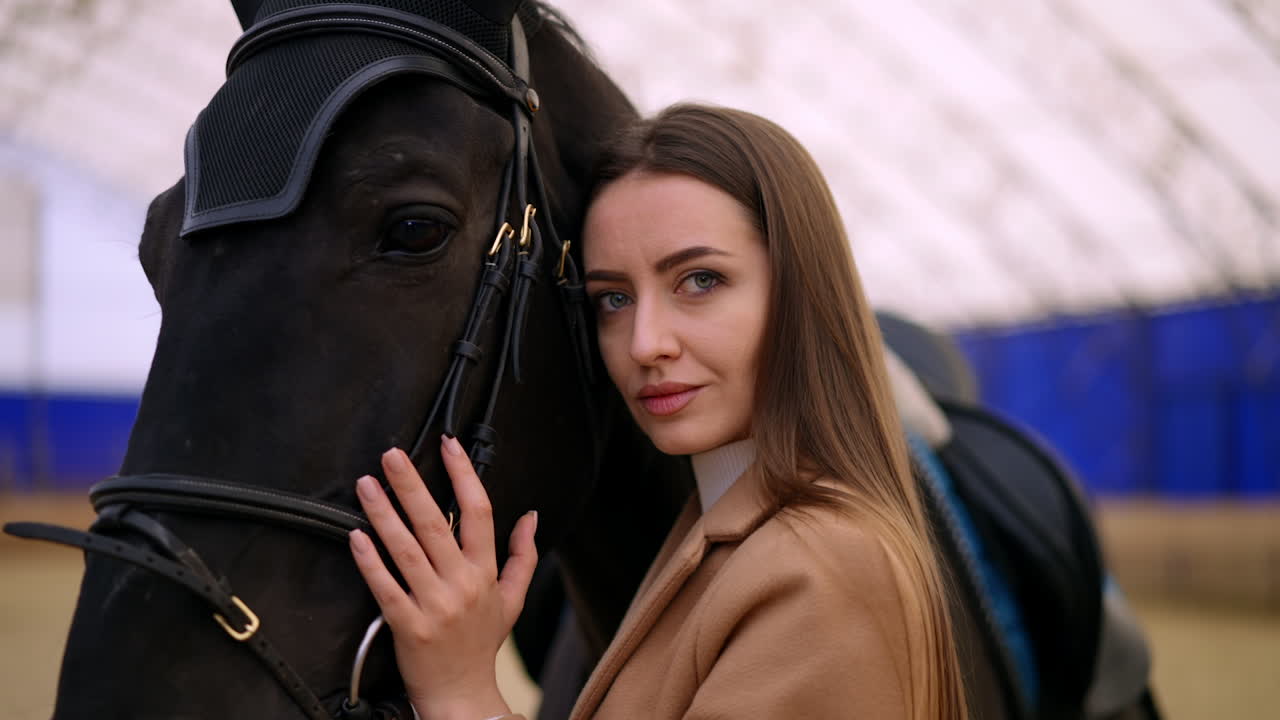 Beautiful big-eyed Caucasian woman leaned to a graceful black horse. Close up. Riding lessons at drill hall. Blurred backdrop.
