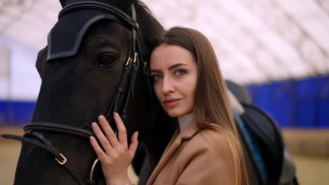 beautiful big-eyed caucasian woman leaned to a graceful black horse. close up. riding lessons at drill hall. blurred backdrop.