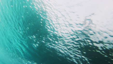 Light-rays-dance-below-ocean-water-surface-as-surfer-catches-wave,-viewed-from-behind