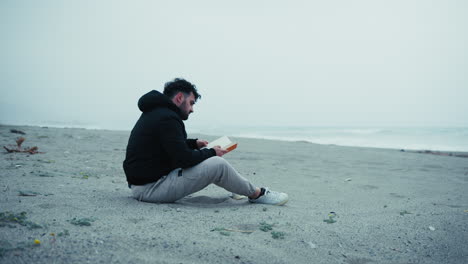 sad man starts reading a book sitting on the sand in winter