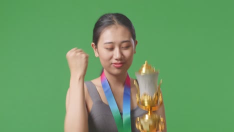 close up of asian woman with a gold medal looking at a gold trophy then celebrating winning as the first winner on green screen background in the studio