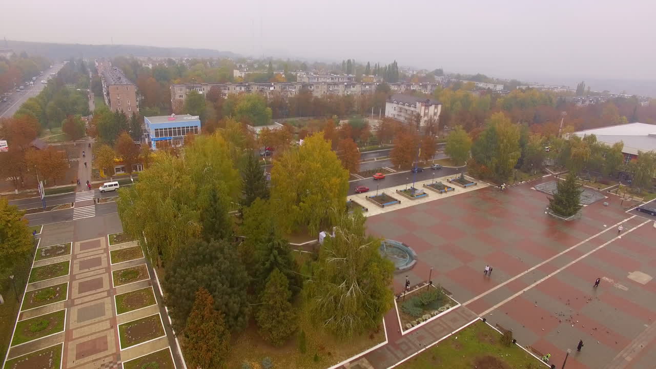 City square in downtown. Trees, fir-trees and flowerbeds around the area. Drone footage going up gradually over the cityscape.