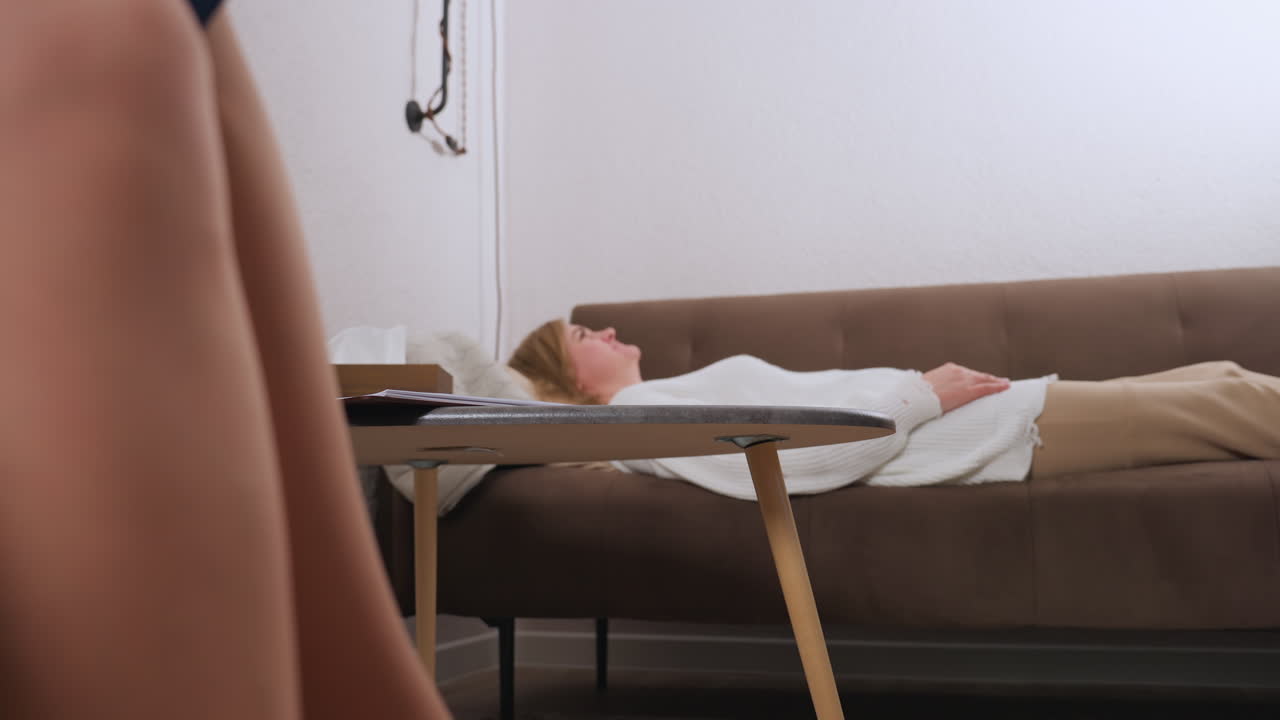 Lower view of mindful guest lying on comfortable long sofa speaking with specialist, tissue box, clock and book on table enhance serene atmosphere during intimate therapy session in quiet room