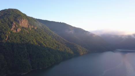 soft light of sunrise shines on rock face hidden in lush green forest beside a calm lake