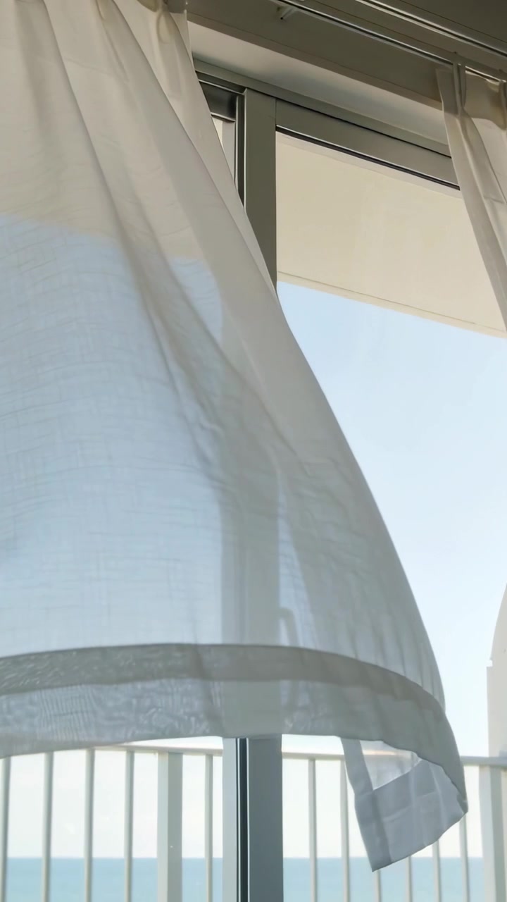 Video verticale: Tenda trasparente che ondeggia rivelando una maniglia e il mare, con la brezza che entra dal balcone.