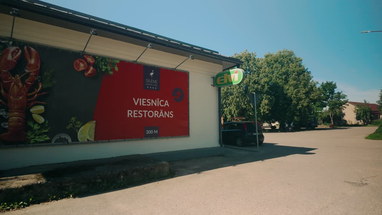 Quiet square in center of Silene, Latvian village near Belarus border, captured on sunny summer day. Includes a large restaurant advertisement and small shop, reflecting the simplicity of rural life.