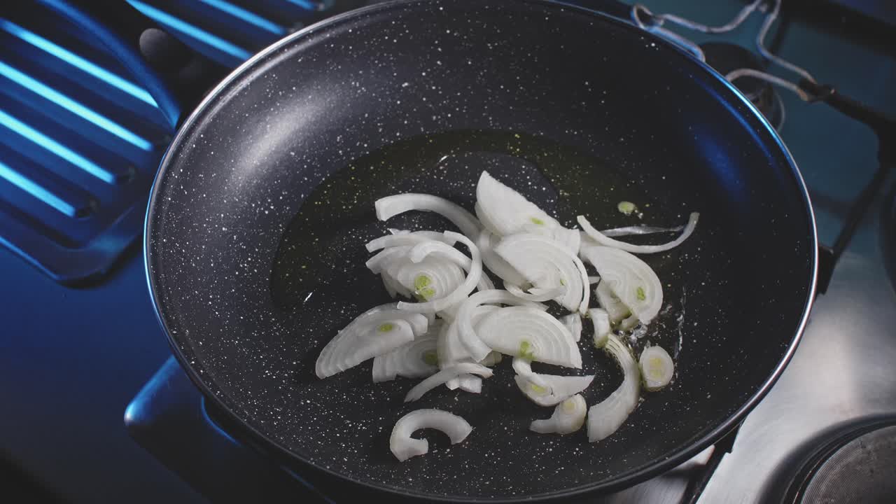 An overhead closeup of a hot oiled skillet where a man's hands transfer sliced onions then adds multicolored bell peppers into the skillet and then stirs with a wooden spatula, spreading evenly