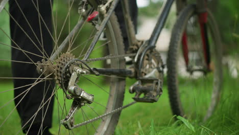 close-up of individual wearing black trousers and sneakers standing beside parked bicycle in grassy field, adjusting the pedal backward, lifting rear tire, and pedaling to rotate the wheel