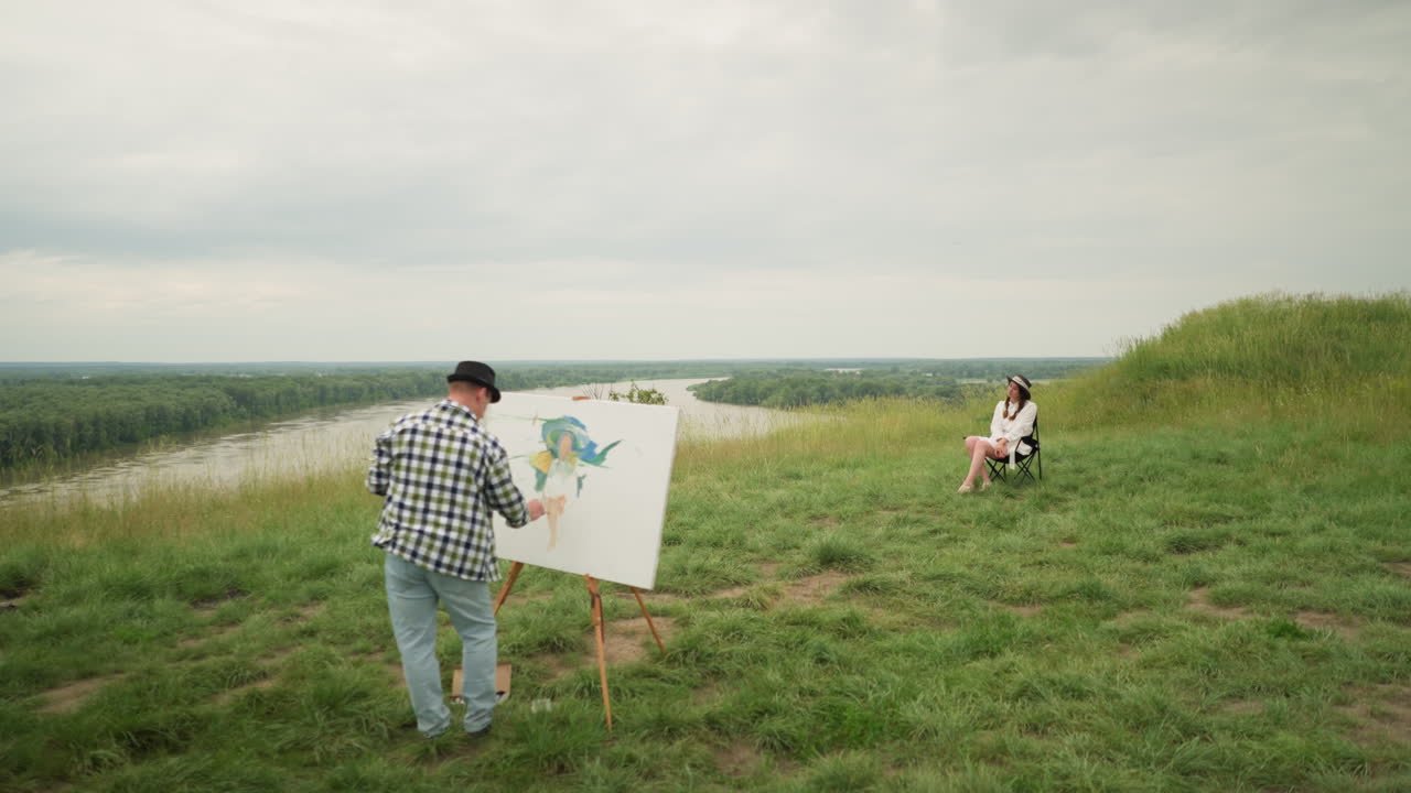 A painter, wearing a hat and checkered shirt, is focused on painting on a board in the middle of a lush grass field under a cloudy sky. a woman in a hat and white dress sits comfortably on a chair