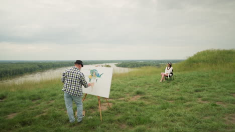 a painter, wearing a hat and checkered shirt, is focused on painting on a board in the middle of a lush grass field under a cloudy sky. a woman in a hat and white dress sits comfortably on a chair