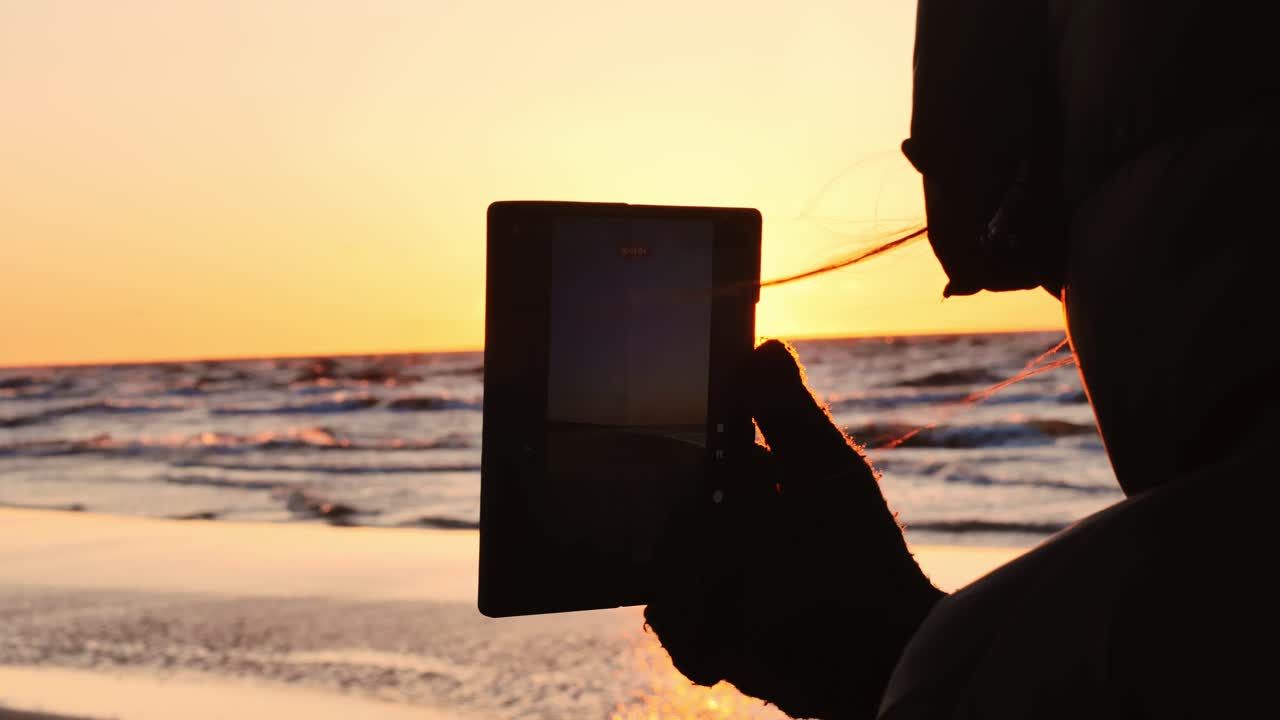 Bright orange sun reflects on tablet screen while person films Baltic Sea sunset