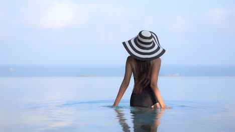 Mujer-Sexy-Con-Sombrero-En-Traje-De-Baño-Negro-De-Una-Pieza-Caminando-Hacia-El-Mar-En-Calma