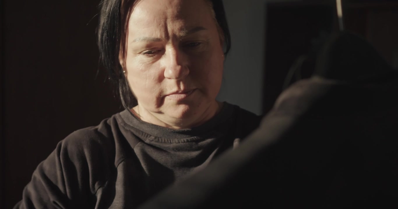 Morning sunlight illuminates a female wardrobe caretaker brushing the black fabric of a traditional miner's uniform beside veiled curtains.
