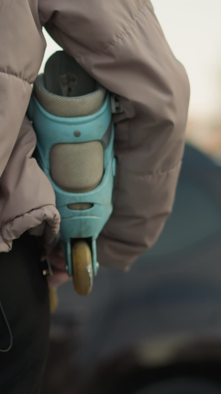 toma de primer plano de una chica con una chaqueta de melocotón y pantalones negros sosteniendo una patineta turquesa, con su largo cabello marrón visible, de pie en un camino del parque mientras un coche se acerca en el fondo