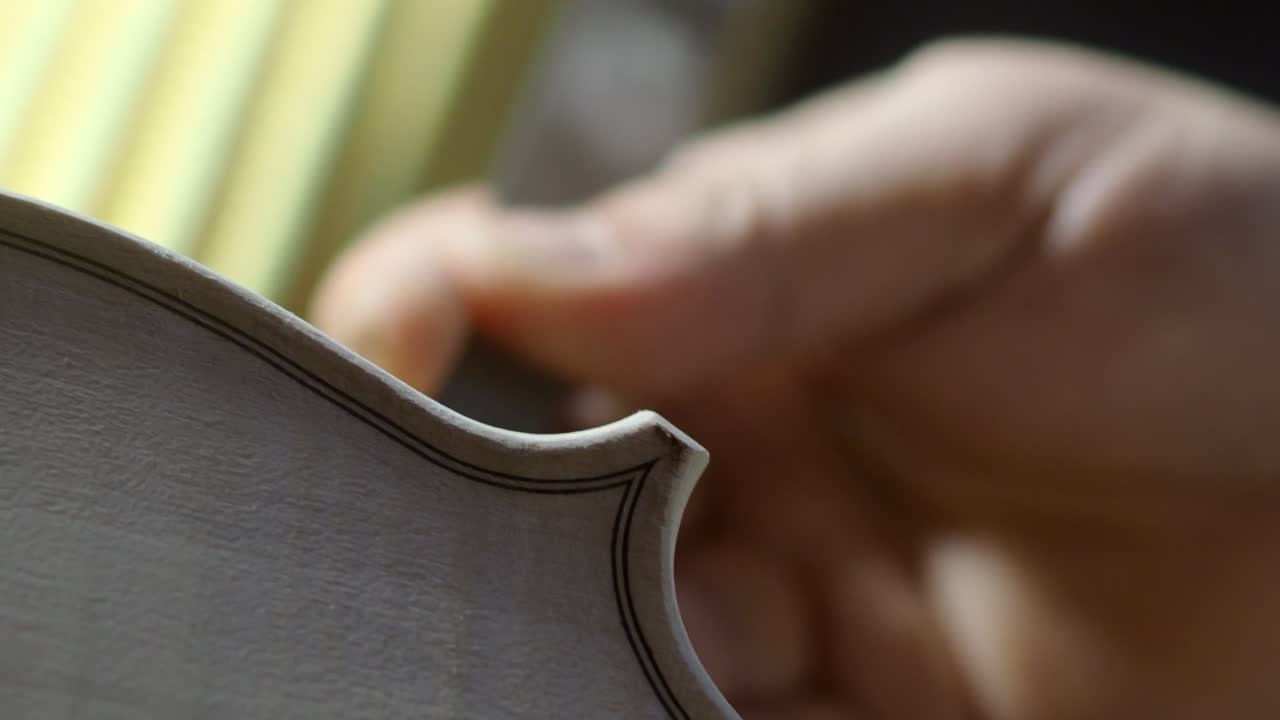 Close-up shot of a luthier’s hand refining the purfling edge on a spruce violin top plate using a fine hand tool, showing careful craftsmanship, tradition, and accuracy in shaping acoustic detail