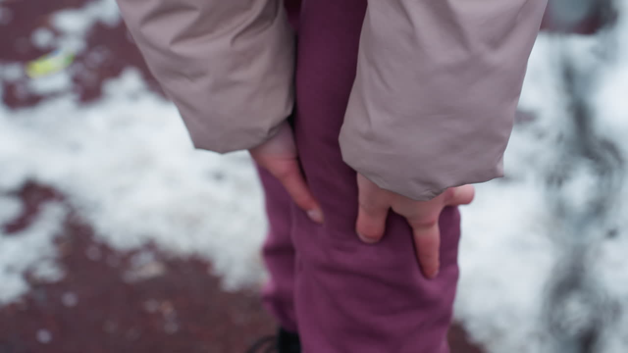 겨울 옷을 입은 사람이 무을 문지르고, 아마도 통증이 있고, 손이 갈색 바지를 고 다리를 잡고, 눈이 흐릿한 배경과 사슬이 근처에 보입니다.
