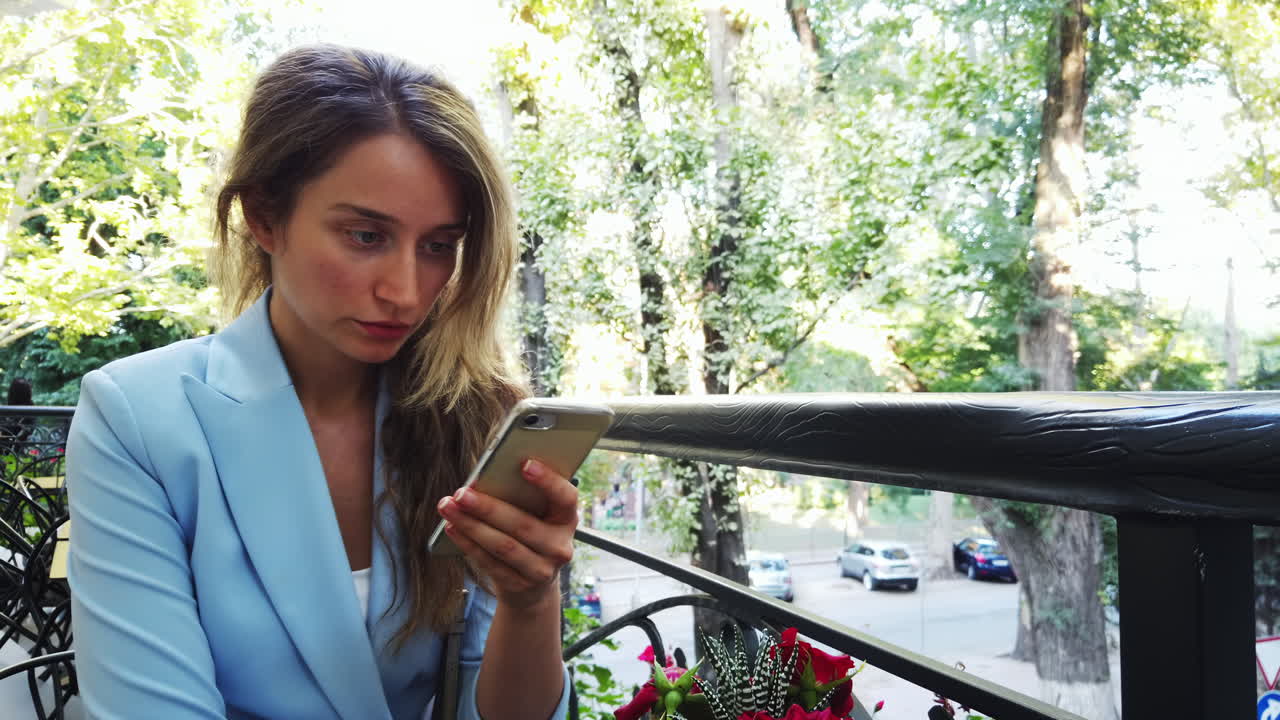 Eine Frau in einer hellblauen Jacke sitzt an einem Café-Tisch und blickt aufmerksam auf ihr Smartphone. Um sie herum sind Bäume und ein Blick auf geparkte Autos an einem sonnigen Tag.