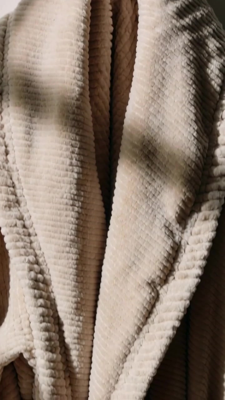 Video verticale: Telecamera che riprende con una panoramica una vestaglia a coste beige appesa a un gancio, mostrando la cintura per il dettaglio del tessuto.