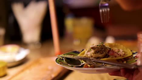A person uses a spoon and fork to pick up a meat taco from a wooden board in a warmly lit Bangkok restaurant, with close-up camera angles