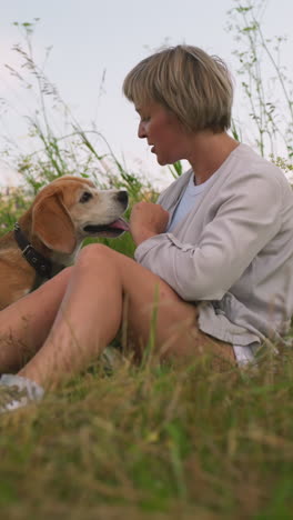 pet lover seated on grassy field with her dogs, playfully kissing one while pushing it back as it gets closer, with leash around their necks, other dog sits nearby watching