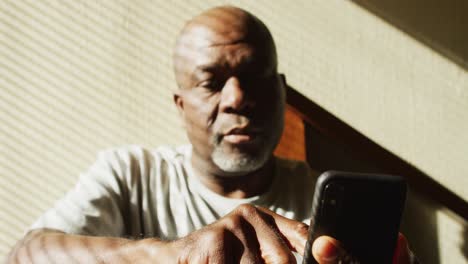 hombre mayor afroamericano pensativo sentado en casa al sol usando un teléfono inteligente