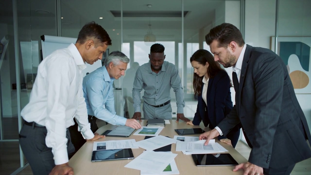 Pointing man in blue shirt moving tablet while team leaning over conference table, scanning reports