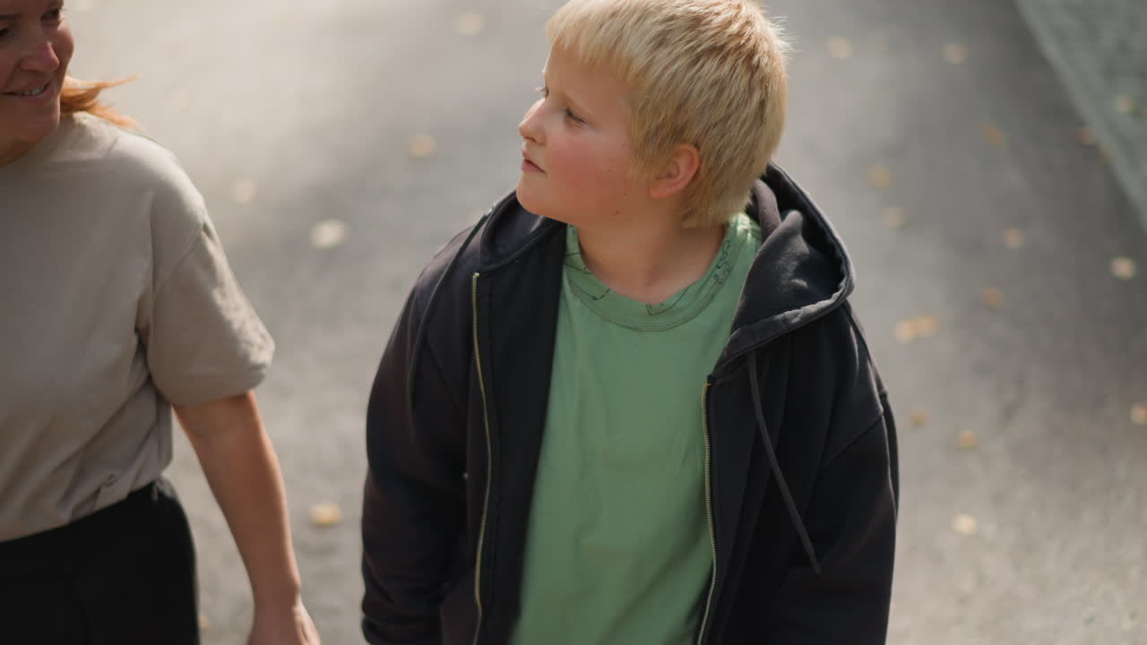 Blonde Boy Looking Up Outdoors Hoodie Green Shirt On Sidewalk Under Soft Sunlight Candid Portrait Of Curious Child Glancing Toward Guardian, Subtle Autumn Leaves On Pavement, Warm Tonal Mood