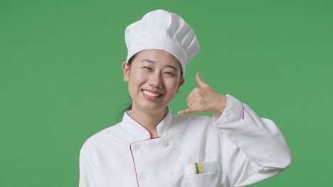 Close Up Of Asian Woman Chef Smiling And Making Call Me Gesture To Camera While Standing In The Green Screen Background Studio