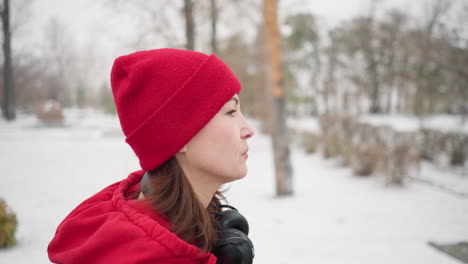 vista lateral de un atleta con gorra roja y chaqueta caminando por un parque nevado con auriculares alrededor del cuello y una expresión intensa, rodeado de árboles desnudos, un paisaje invernal sereno