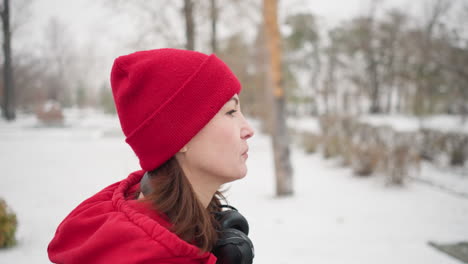 side view of athlete wearing red beanie and jacket walking through snowy park with headphones around neck and an intense expression, surrounded by bare trees, serene winter scenery