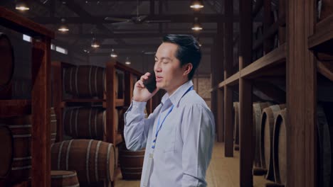 side view asian male professional worker standing in cellar of winery, he pick the phone up and started to specking to it with a slicely smile