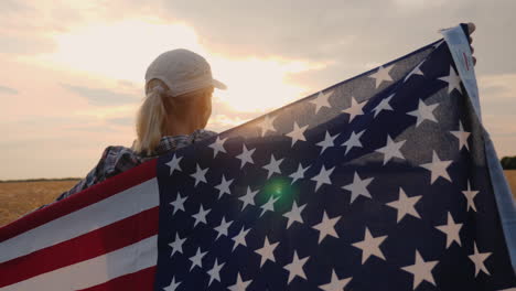 A-Woman-Farmer-With-The-Usa-Flag-Stands-In-A-Field-Of-Ripe-Wheat-At-Sunset-Independence-Day-Concept