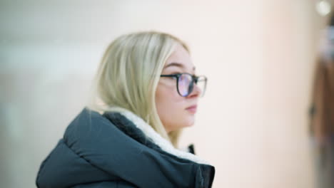 young lady with glasses walking past well-lit clothing store in mall, clothes displayed on mannequin visible through glass with soft bokeh light effect in blurred background