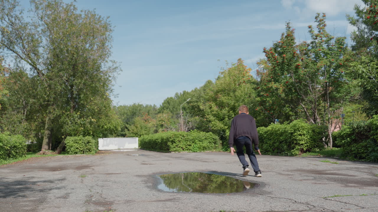 Uomo bianco in equilibrio su un sentiero di ghiaia, braccia tese fingendo di volare, che cammina verso una pozzanghera sotto un cielo blu, giacca casual, alberi verdi e arbusti che incorniciano uno spazio aperto, movimento giocoso all'aperto