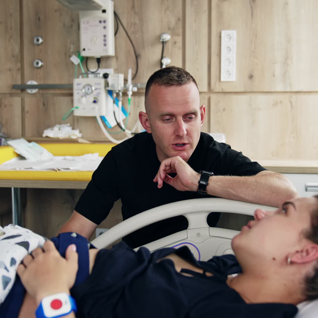 Man wearing black t-shirt talking to his wife lying in hospital bed. Woman is checked before baby delivery