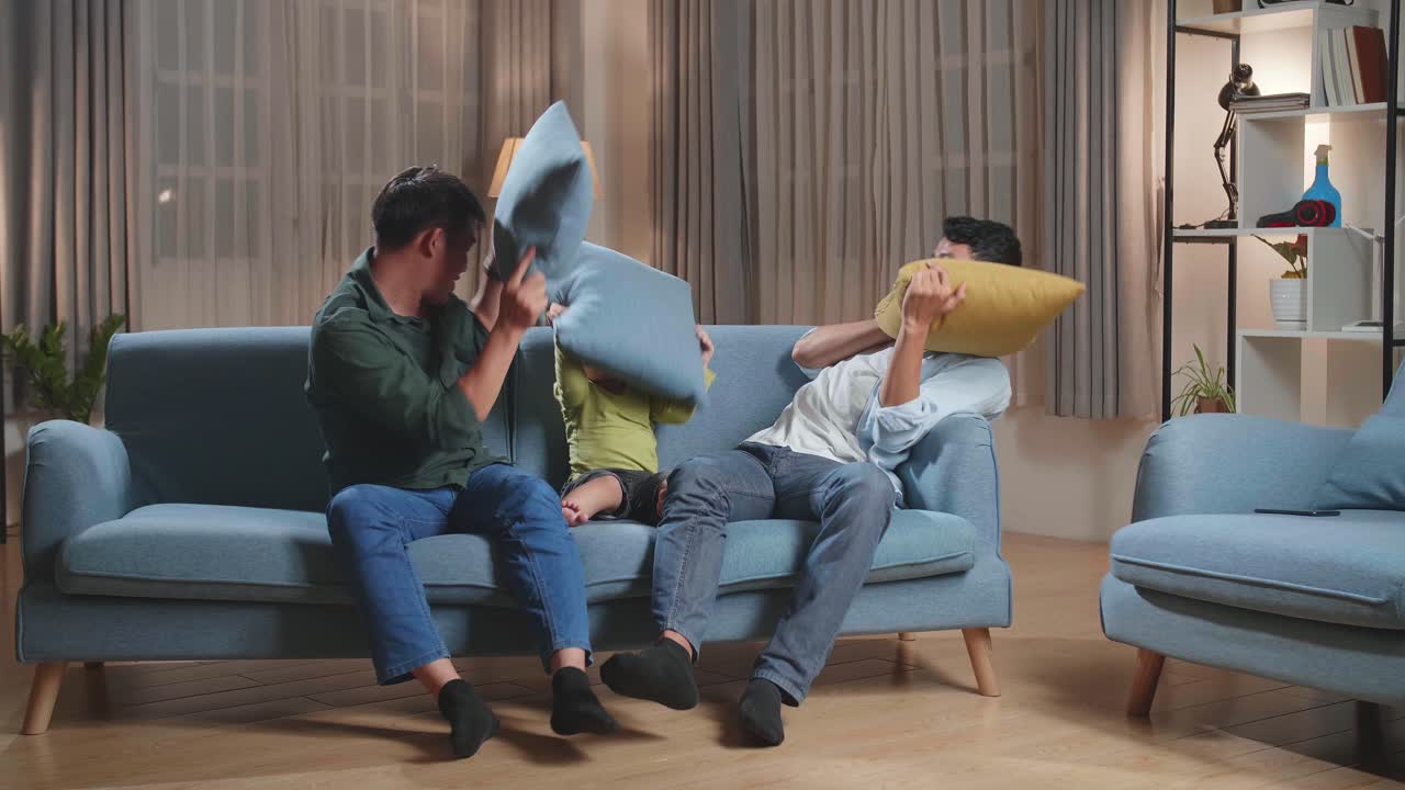 Happy Asian Gay Couple Having Fun With Their Lovely Son Fighting Pillows In Living Room