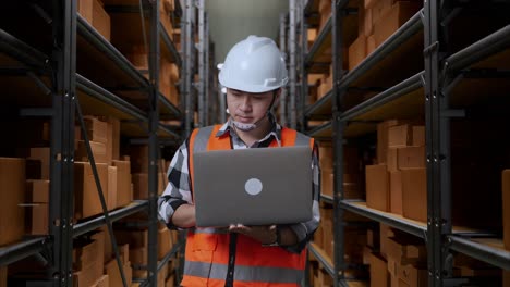 asian male engineer with safety helmet looking at a laptop and looking around while standing in the warehouse with shelves full of delivery goods