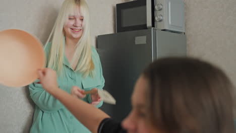 two young women in cozy kitchen use mixing bowl and spoon as playful props tapping and dodging utensils around potted plant and snack bag creating lighthearted joyful sibling fun moment