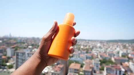 persona sosteniendo una botella de protector solar con una ciudad en el fondo