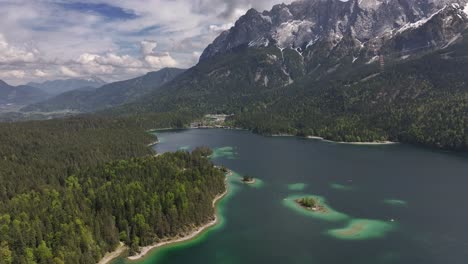 Vista-Aérea-Del-Lago-Eibsee-Rodeado-De-Exuberantes-Bosques-Verdes-Y-Majestuosas-Montañas-Cubiertas-De-Nieve-Bajo-Un-Cielo-Parcialmente-Nublado-En-Grainau,-Alemania