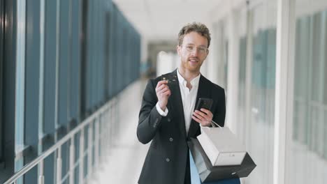 portrait of adult stylish man with paper bags showing smartphone and credit card for shopping