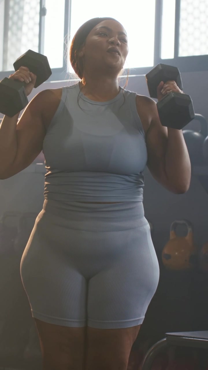 Video verticale: Donna che solleva manubri in palestra per le spalle, indossando un top grigio e pantaloncini.