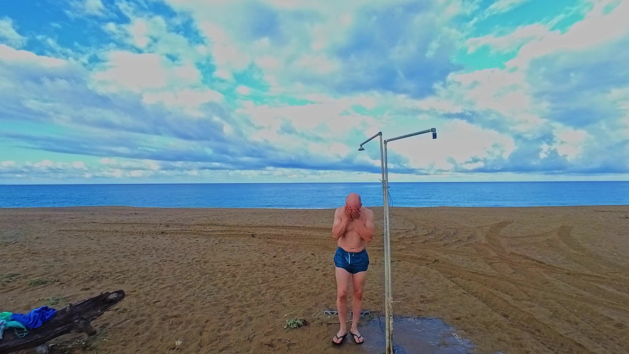 Inquadratura ampia di un uomo che si sciacqua con acqua dolce sotto una doccia da spiaggia all'aperto dopo aver nuotato, poi si allontana per asciugarsi con un asciugamano contro un orizzonte oceanico nuvoloso