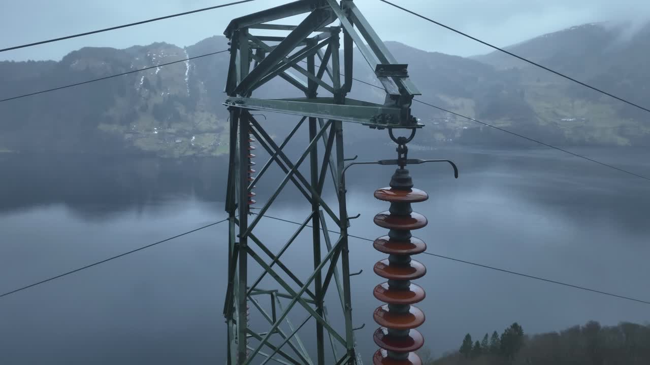 High voltage pylon top with insulators in focus. Drone descends slowly while tilting up, creating parallax against a fjord background in rainy, misty weather