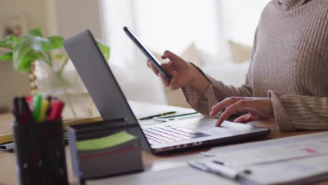 video of midsection of biracial woman at home sitting at desk working on laptop and using smartphone