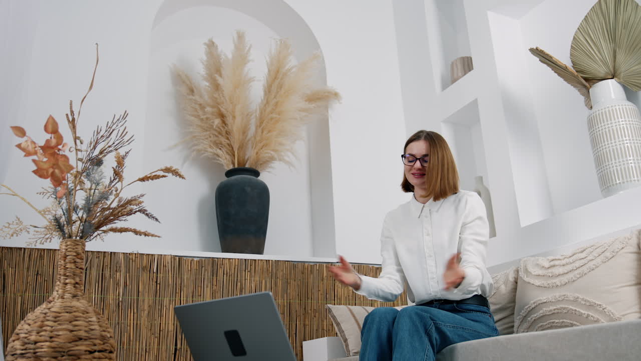 Smiling young woman sits on sofa looking at laptop on the coffee table. Lady is having online video chat via internet.