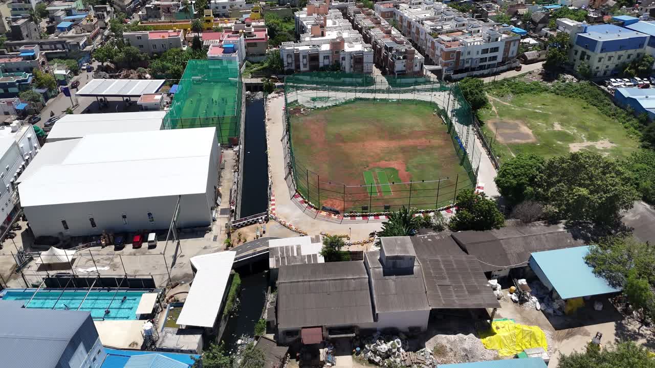 Aerial footage of Sports Field cricket ground Towards the top middle, there's a sports field that looks like a baseball or softball diamond, surrounded by netting or a low fence