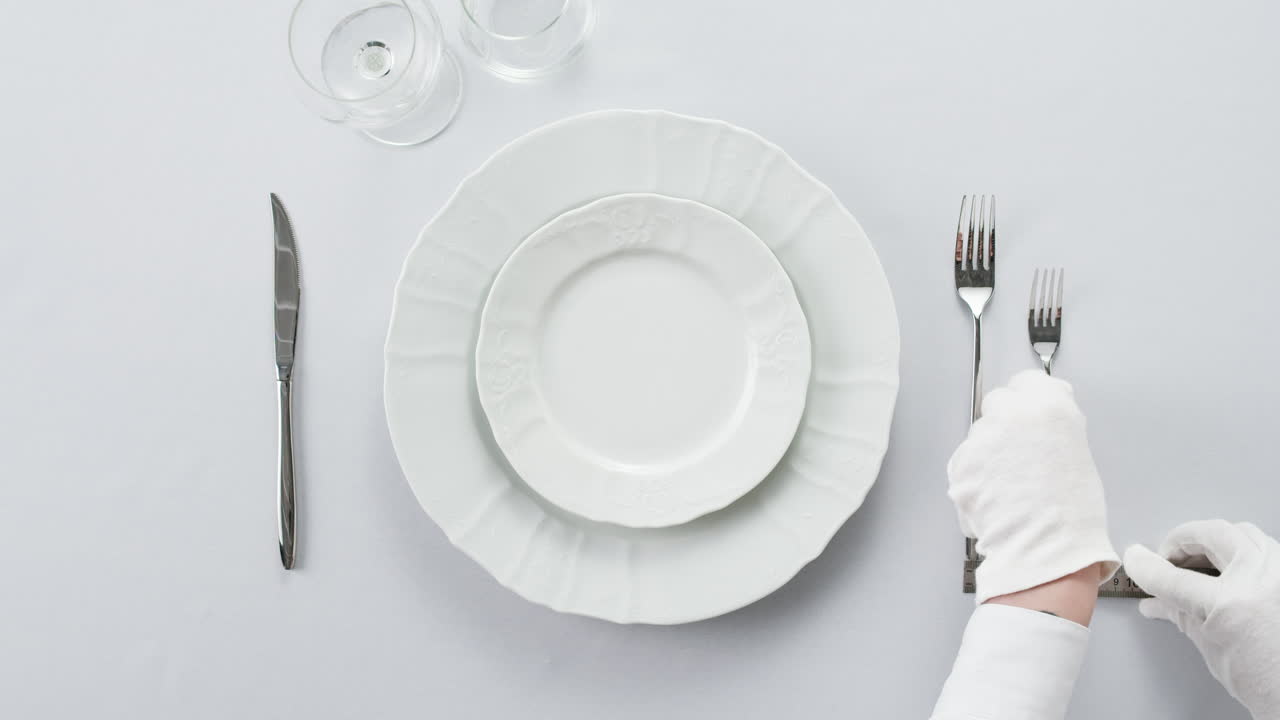 Professional table setting with white gloves. Plates, fork, knife, and glass arranged on a white table.