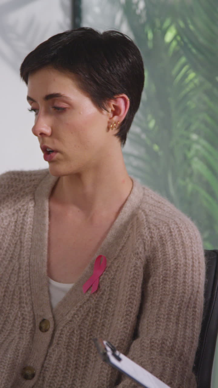 Vertical Video Of Young Woman Wearing Pink Breast Cancer Awareness Ribbon Meeting And Talking At Therapy Support Group For Cancer Treatment Patients With Counsellor